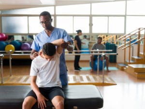 male healthcare provider stretching the arm of a teenage boy sitting on a massage table during physical therapy session