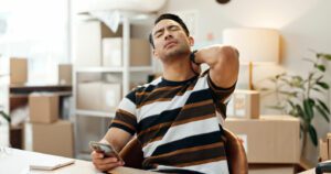 A man is suffering from chronic neck pain while using his phone at home.