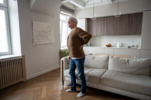 An elderly man grimaces as he slowly straightens his hunched back after standing up from a couch, experiencing severe spinal pain associated with spinal stenosis.